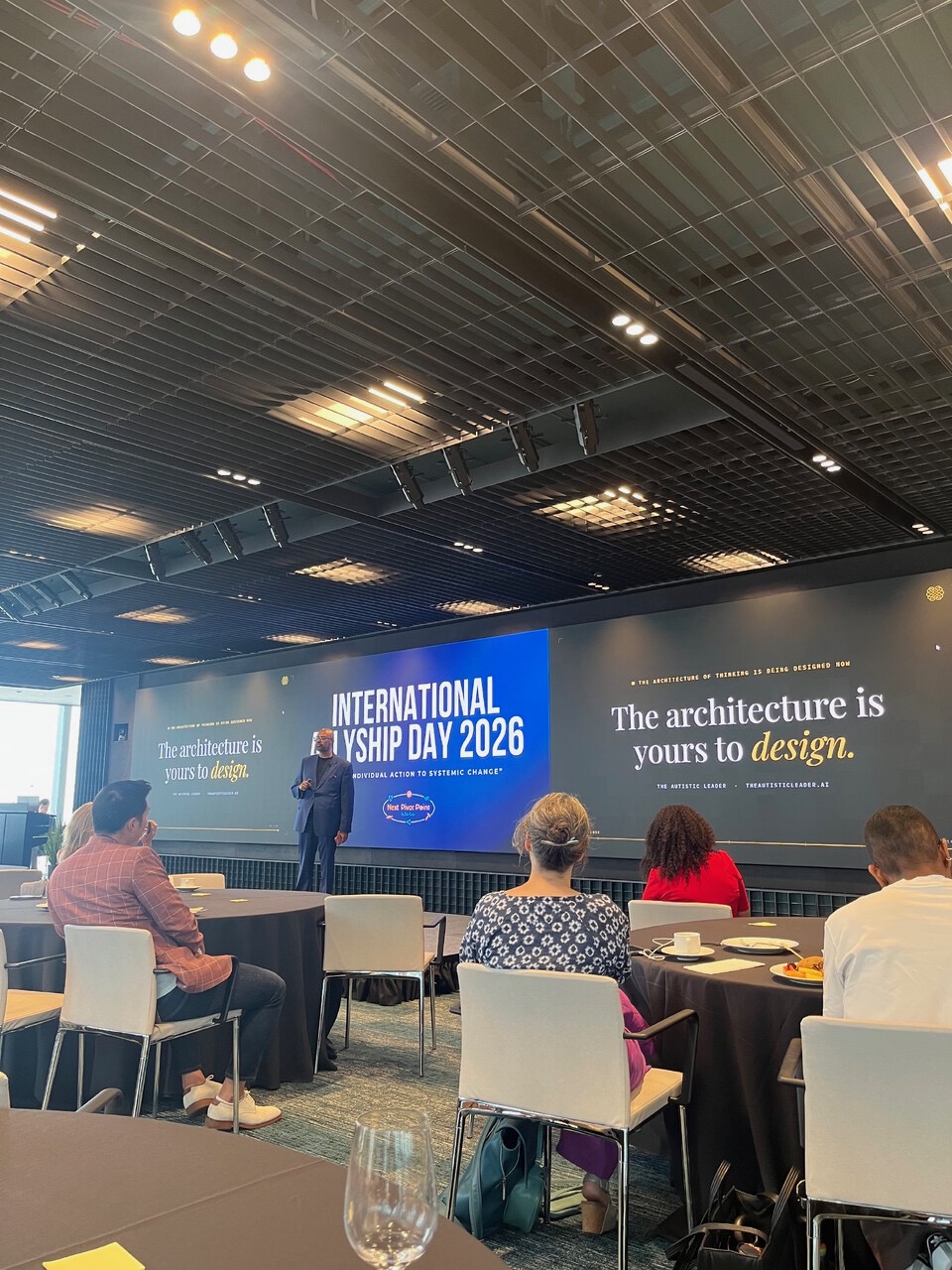 Uday Kiran Bolusani delivering The Undesigned Mind keynote at International Allyship Day 2026 in New York City. The stage screens read 'The architecture is yours to design.' beside the International Allyship Day 2026 title card.
